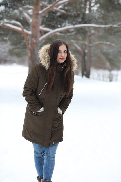 A woman standing in a snowy landscape, wearing a long, hooded parka with a fur-lined hood. The parka is a dark green color, and she is wearing snow boots and gloves.