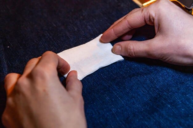 A close-up shot of hands sewing a patch onto a denim jacket. The patch is colorful and floral, adding a personal touch to the garment. The background is a cluttered but cozy craft room with various fabrics and sewing supplies.
