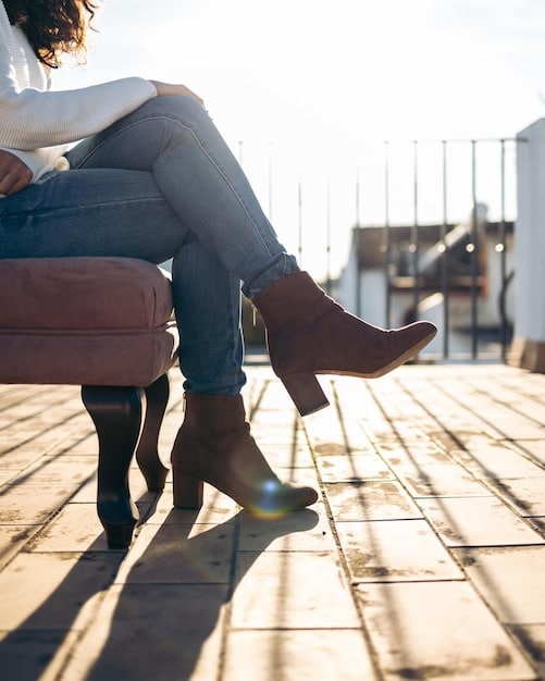 A close-up of a woman's feet wearing stylish brown leather ankle boots paired with dark wash skinny jeans. The background shows a blurred urban setting.