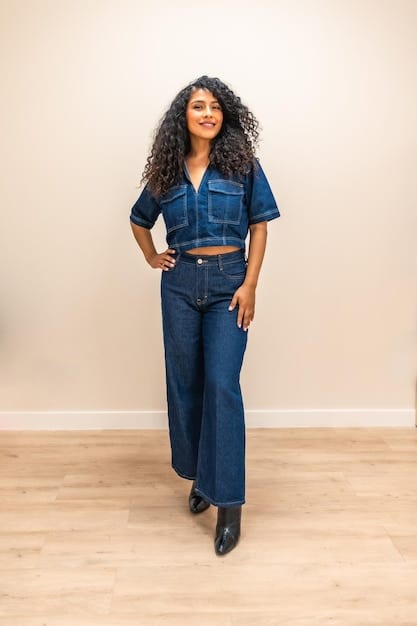 A woman with a curvy figure is wearing a dark wash denim jumpsuit with a wide belt cinching her waist. She is smiling confidently and wearing stylish ankle boots.
