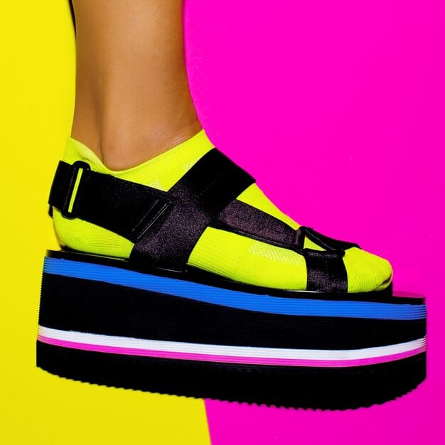 A person wearing chunky platform sandals in a bright, solid color (e.g., yellow or pink) paired with a flowy midi dress. The setting is an urban street with a colorful mural in the background, showcasing the sandals as a fun and stylish statement piece.